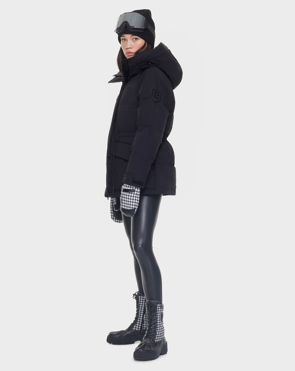 A person wearing a Rudsak MANDORA - 8121542 BLACK women’s down parka, black beanie, ski goggles, black leggings, black-and-white mittens, and black boots stands sideways against a white background.