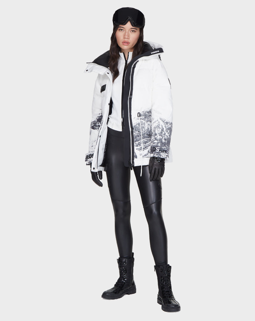 A woman stands facing forward against a plain background, wearing the Rudsak MANDORA - 8121542 MIX women’s down parka with black mountain print, paired with a black beanie, ski goggles, gloves, faux leather leggings, and lace-up boots.