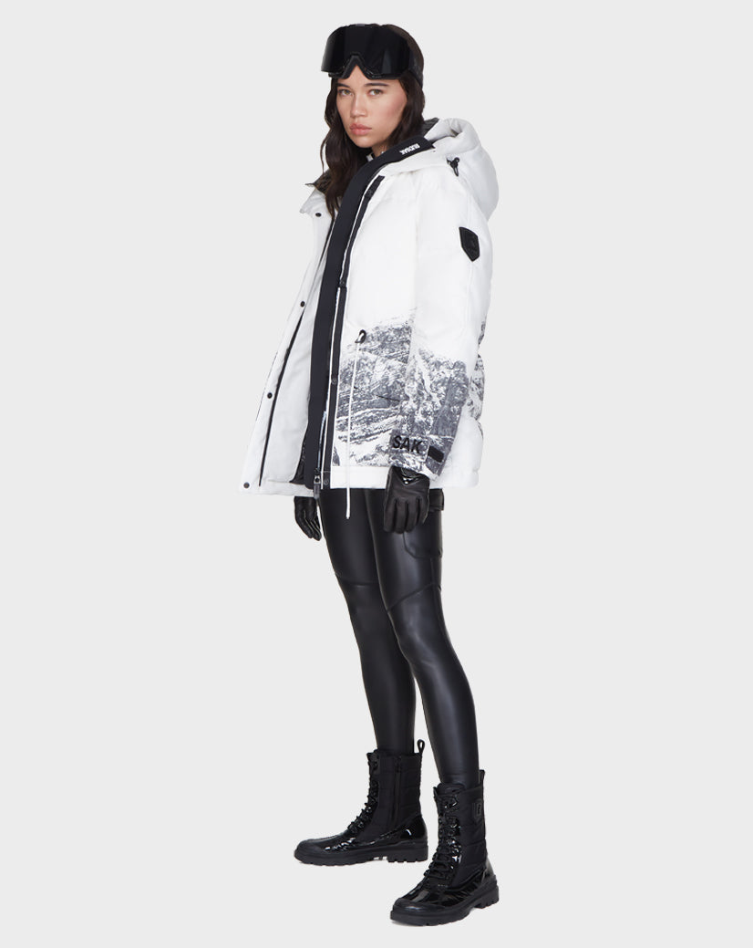 A woman stands slightly sideways against a plain background wearing the Rudsak MANDORA - 8121542 MIX white ski parka with black and white mountain print, paired with black leggings, boots, gloves, and a black ski headband.
