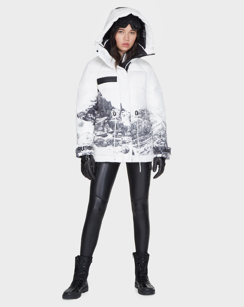 A woman stands facing forward wearing the Rudsak MANDORA - 8121542 MIX white hooded women’s down parka with a black mountain print, paired with black gloves, ski goggles, faux leather pants, and lace-up boots.
