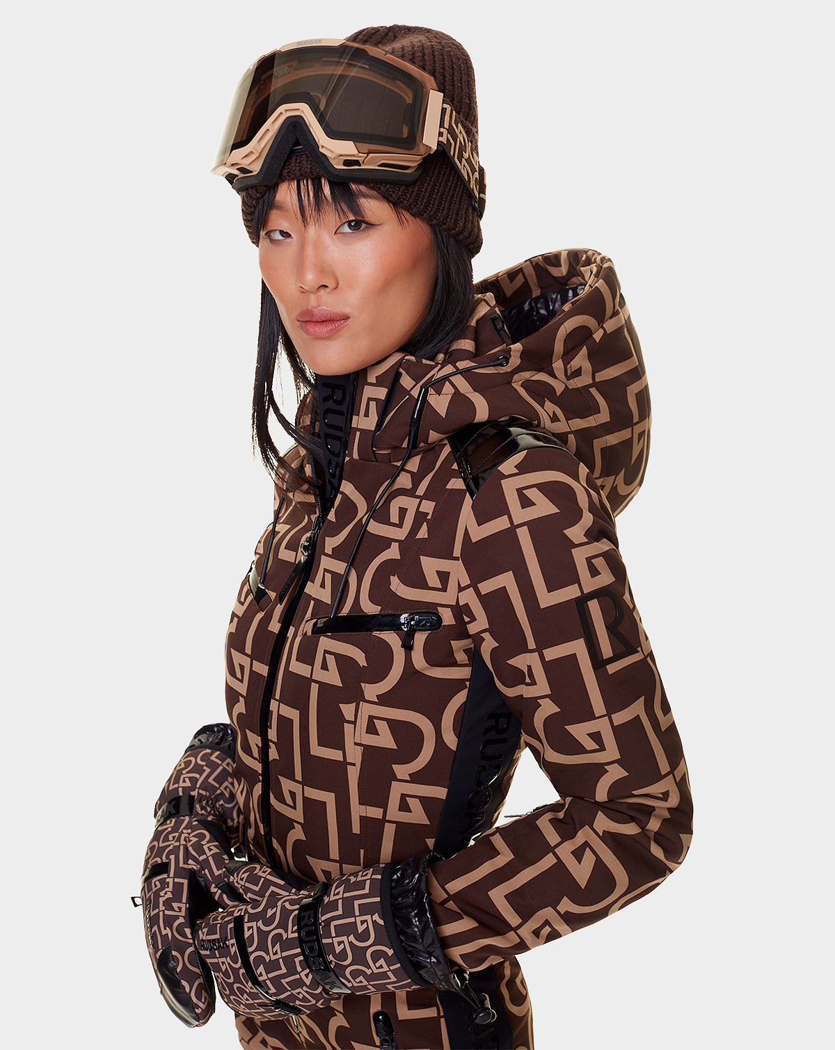 A woman in a Rudsak brown patterned ski jacket and gloves, matching beanie, and tan PEAK - 9421515 RUDSAK PATTERN ski goggles with interchangeable lenses stands against a plain white background.