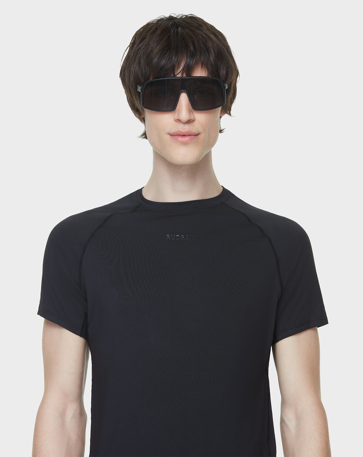 A young person with short brown hair wears Rudsak AKSEL - 9422031 BLACK sunglasses featuring UV Block Lenses and a fitted black t-shirt, standing against a plain white background.
