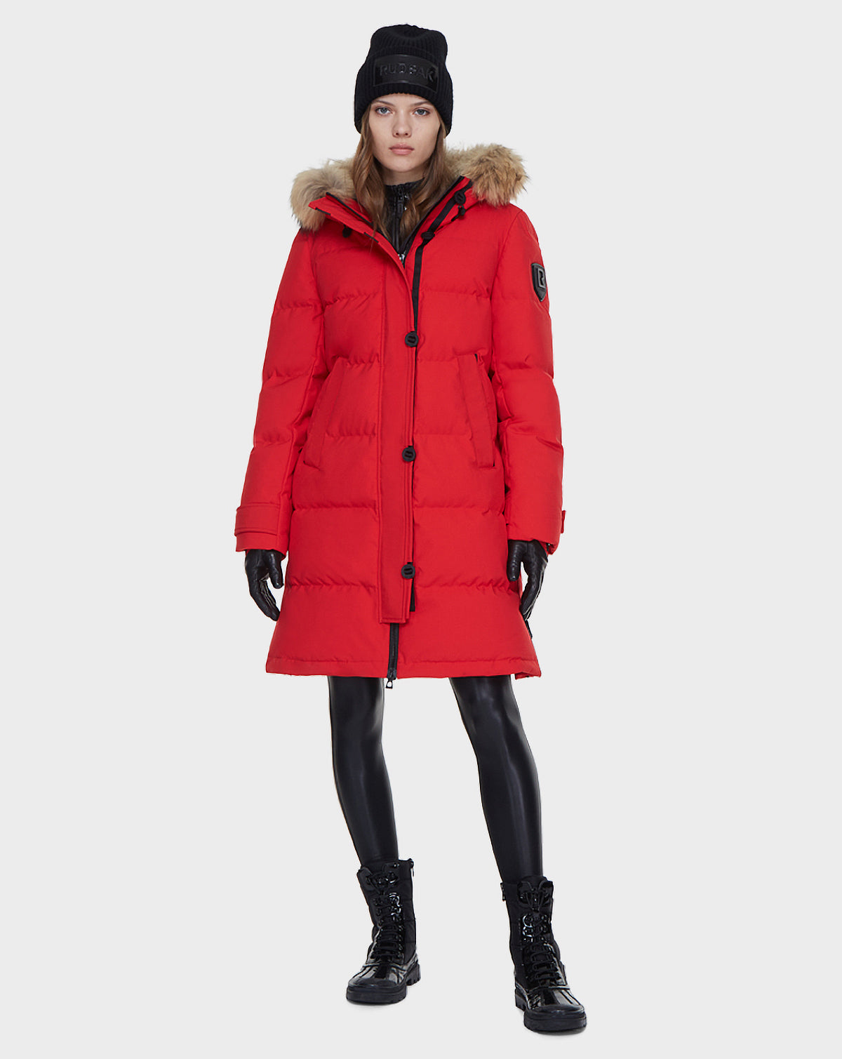 A person stands facing forward against a plain light background, wearing the Rudsak MEYA - L121523 POPPY RED women's down parka with fur-lined hood, paired with black beanie, gloves, leggings, and lace-up boots.
