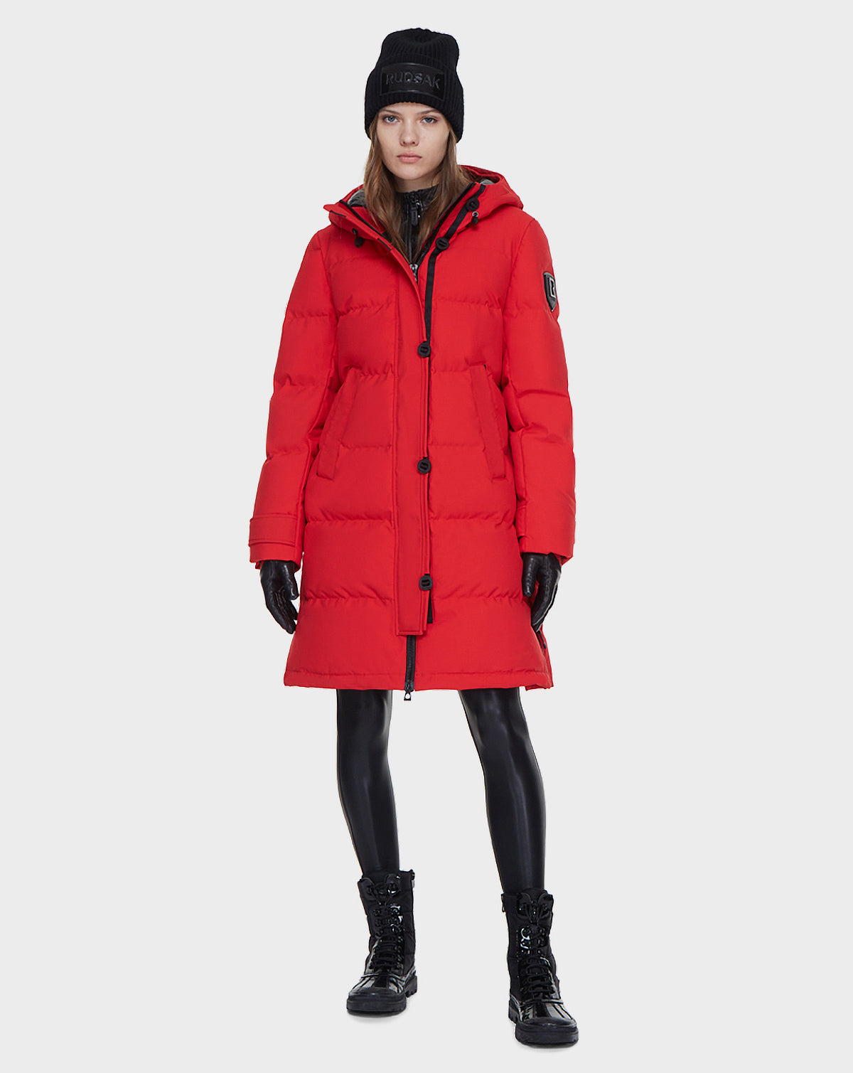 A woman stands facing forward wearing the Rudsak MEYA - L121523 POPPY RED women’s down parka, along with a black beanie, black gloves, shiny black leggings, and lace-up boots against a plain white background.