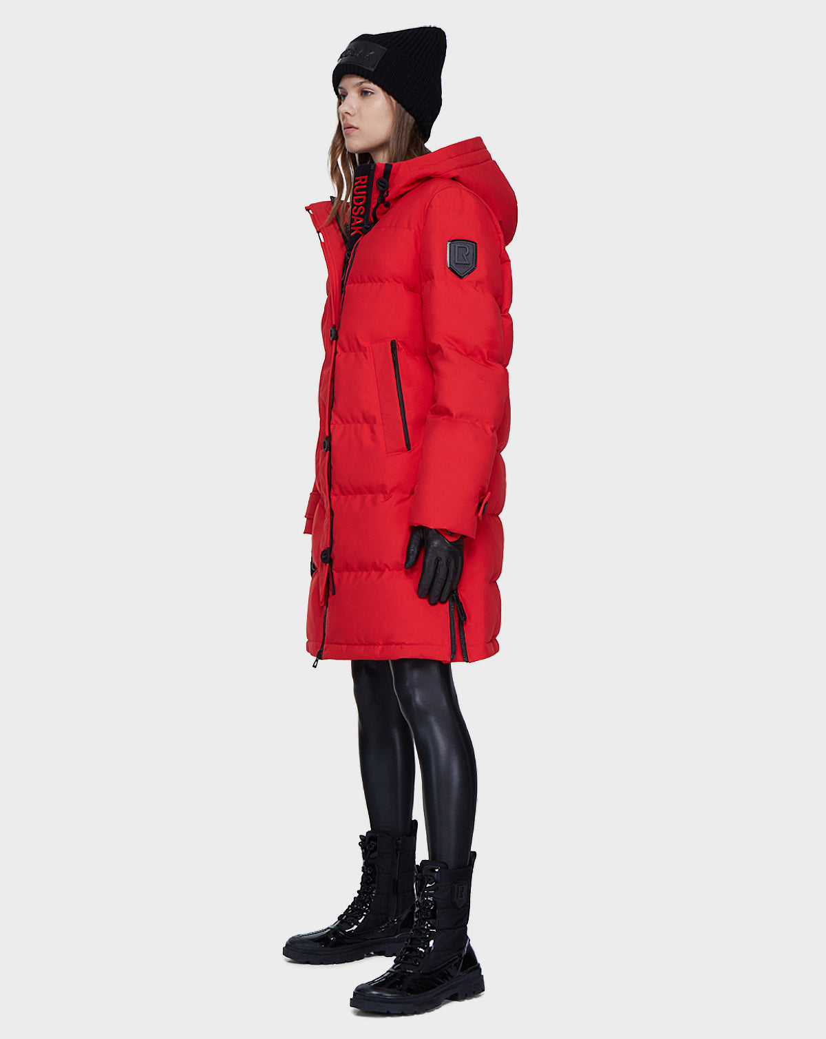 A person wears the Rudsak MEYA - L121523 POPPY RED water-resistant winter coat with hood, paired with a black beanie, gloves, shiny leggings, and boots, standing against a plain light gray background.