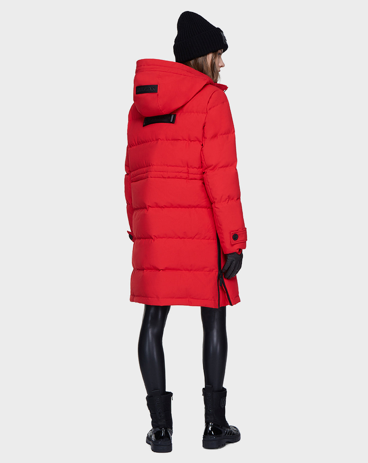 A person wearing the Rudsak MEYA - L121523 POPPY RED women’s down parka, with black beanie, gloves, leggings, and boots stands facing away against a plain light background.