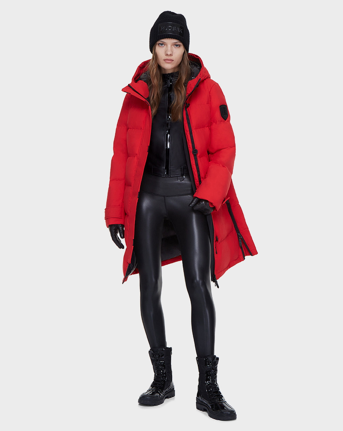 A person faces forward against a plain light grey background, wearing the Rudsak MEYA - L121523 POPPY RED women’s down parka over a black shiny outfit, with black boots, gloves, and a black beanie.