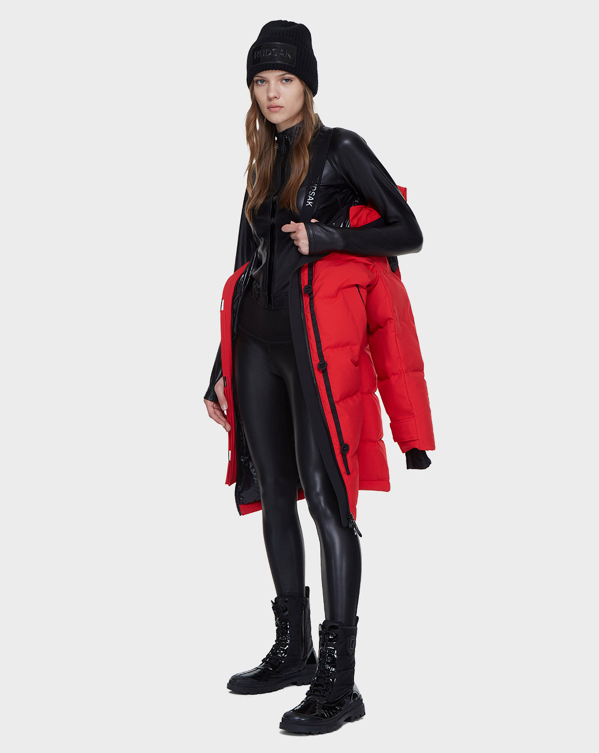 A woman in a black beanie, shiny bodysuit, and lace-up boots stands holding a Rudsak MEYA - L121523 POPPY RED women’s down parka over her shoulder against a plain white background.