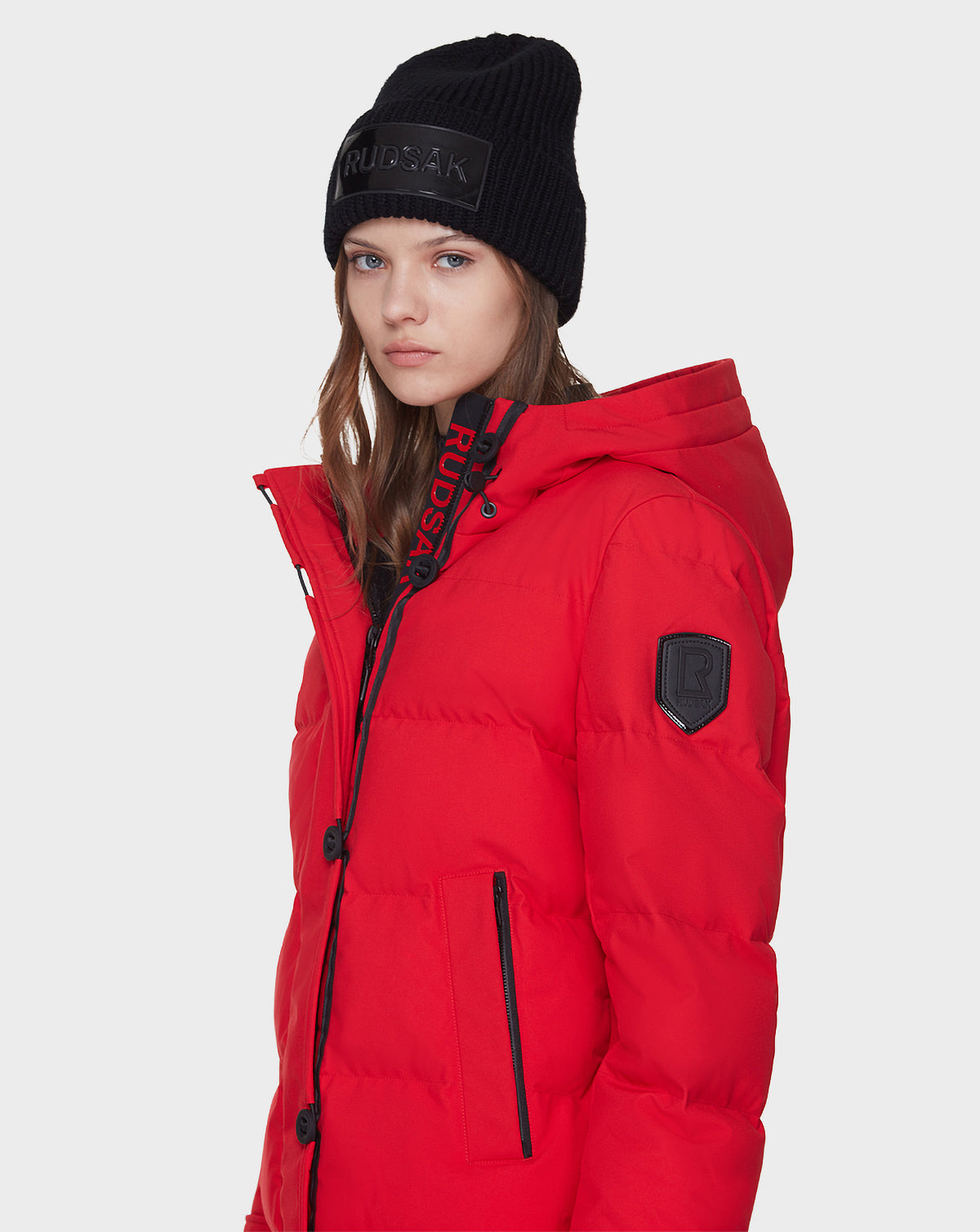 A young woman wears the Rudsak MEYA - L121523 POPPY RED women’s down parka and a black knit beanie, featuring black zipper details and the RUDSAK logo, as she stands against a plain light background looking at the camera.