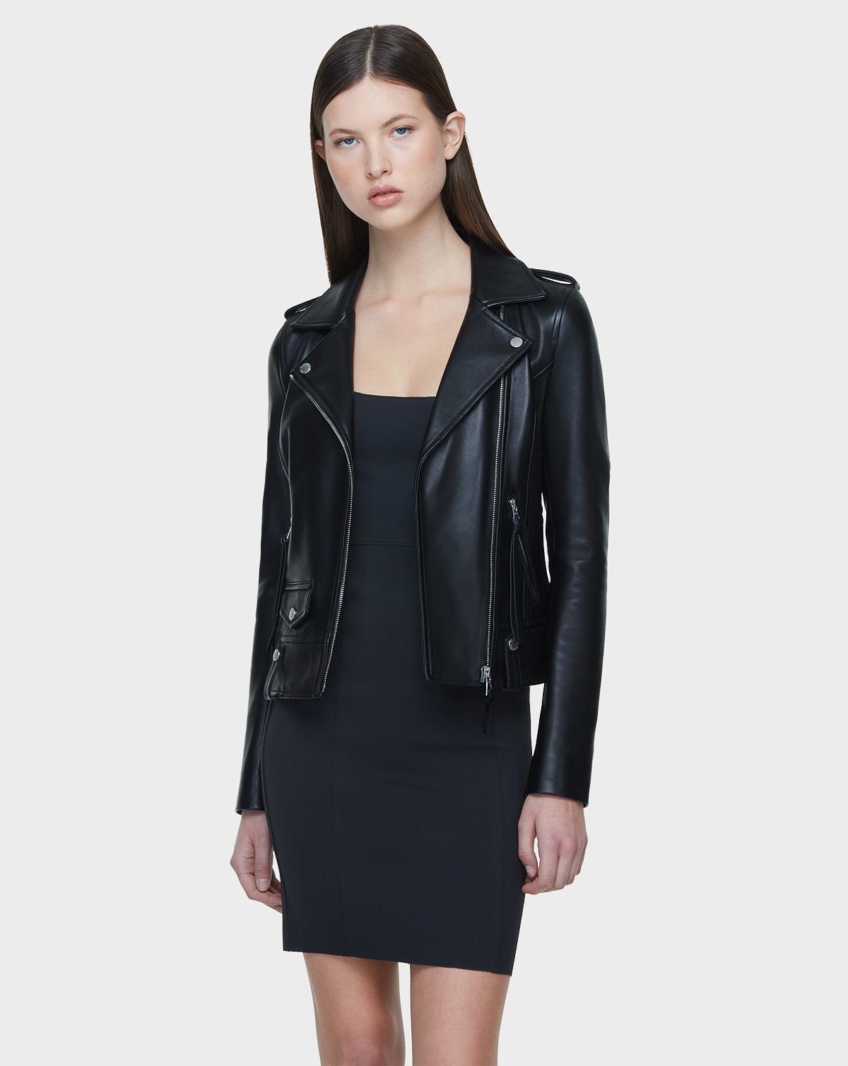 A woman with straight dark hair models the Rudsak MERGO - CA EN Black 8118525 biker jacket, featuring an asymmetrical zip, over a fitted black dress against a plain light gray background.