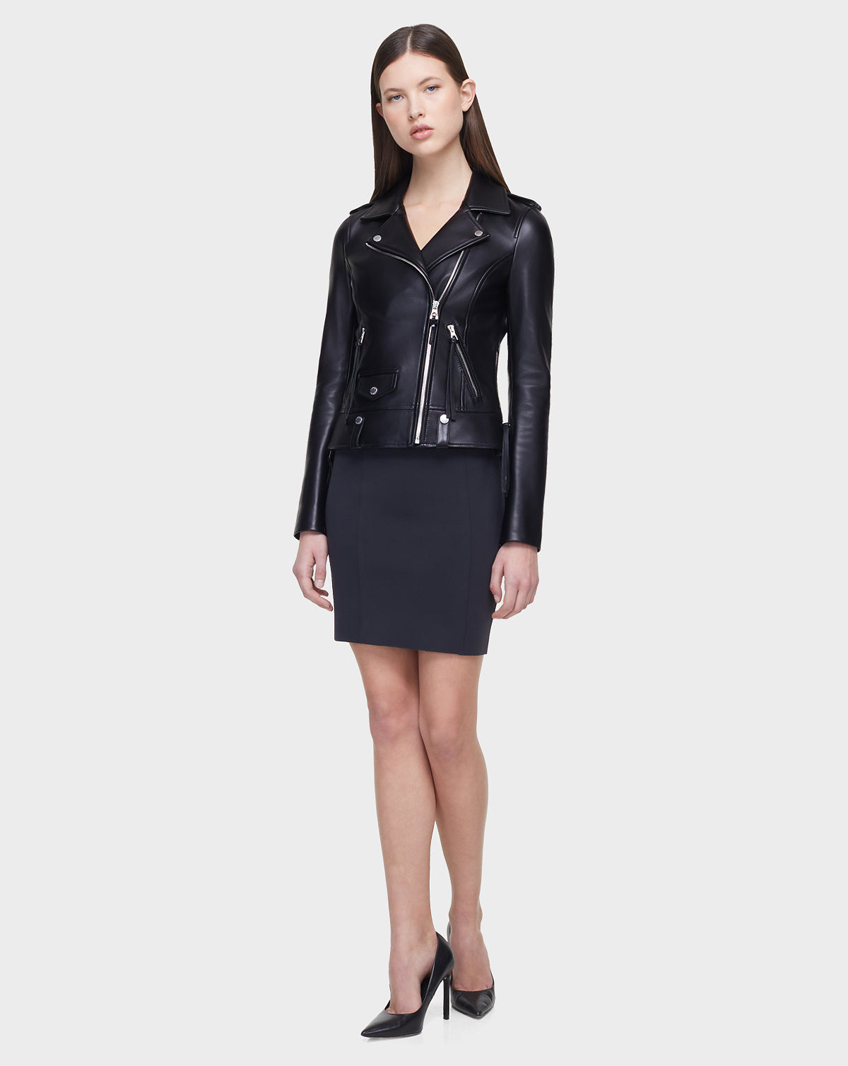 A woman faces forward against a plain light background, wearing the Rudsak MERGO - CA EN Black 8118525 biker jacket with an asymmetrical zip, a dark knee-length skirt, and black high-heeled shoes.
