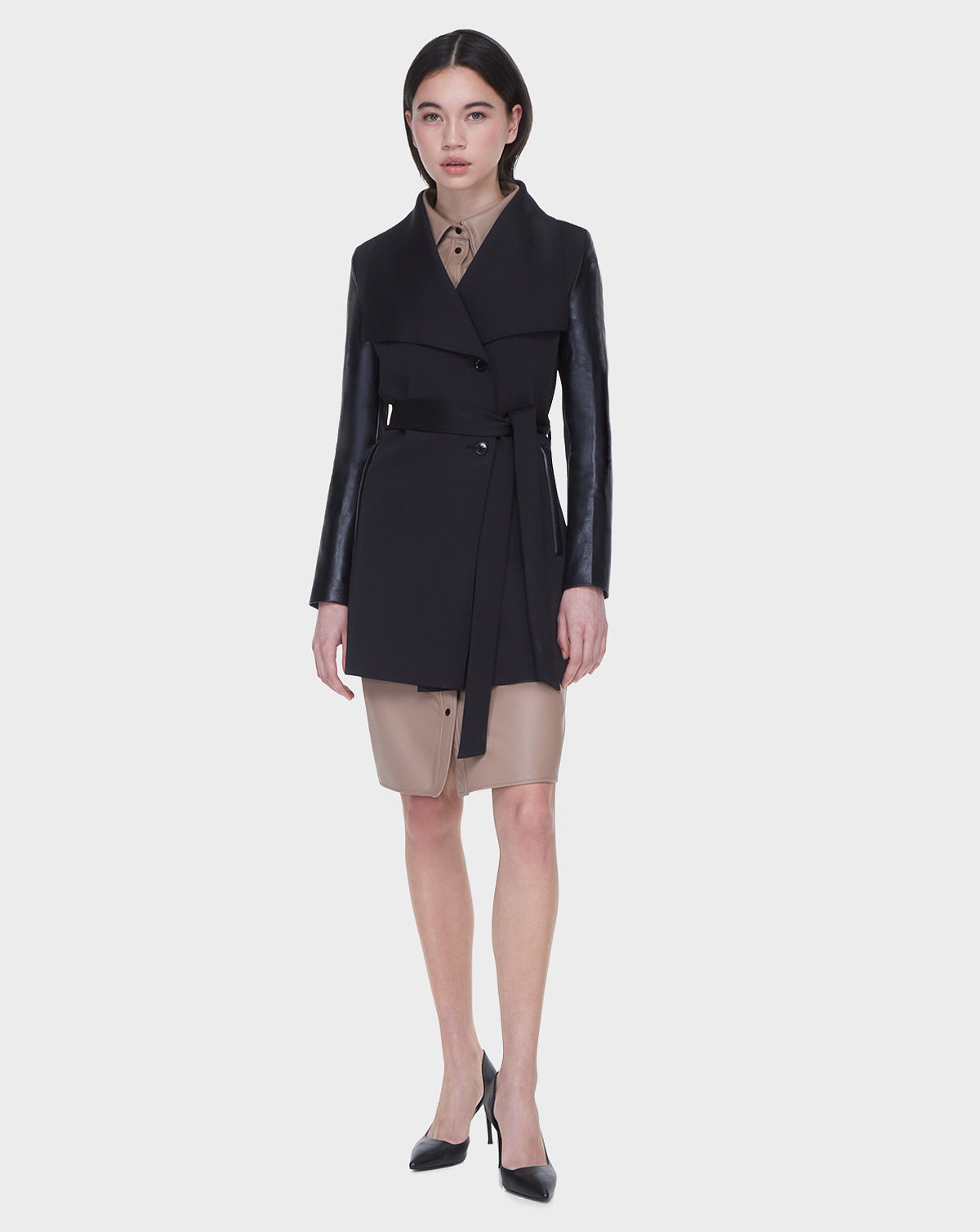 A woman faces forward, wearing the Rudsak MELBA - L122007 BLACK coat with recycled leather sleeves over a tan button-down dress. She has straight dark hair, black pointed heels, and stands against a plain light gray background.