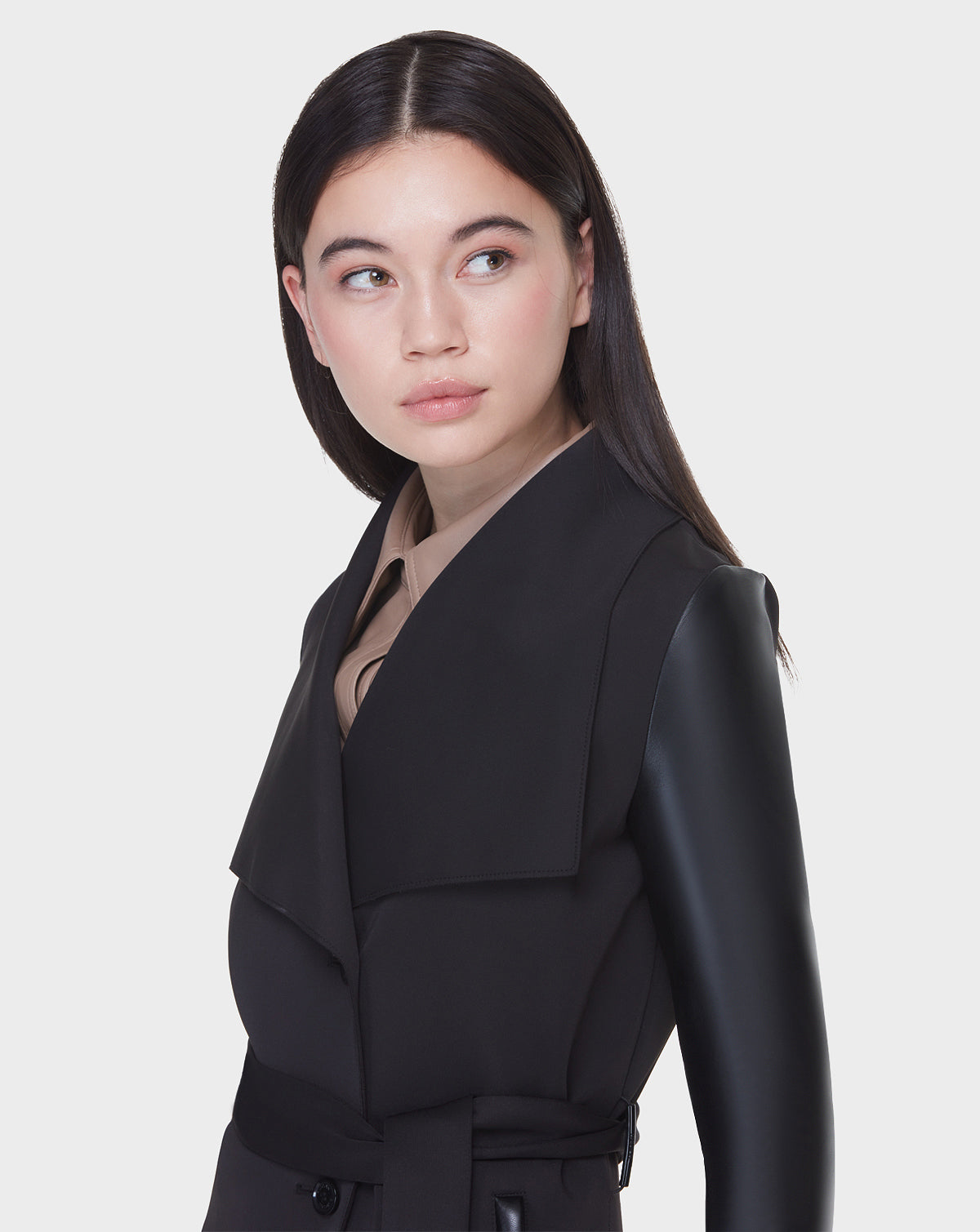 A woman with straight dark hair wears the Rudsak MELBA - L122007 BLACK women's coat with wide lapels and recycled leather sleeves, looking to her left against a plain white background.