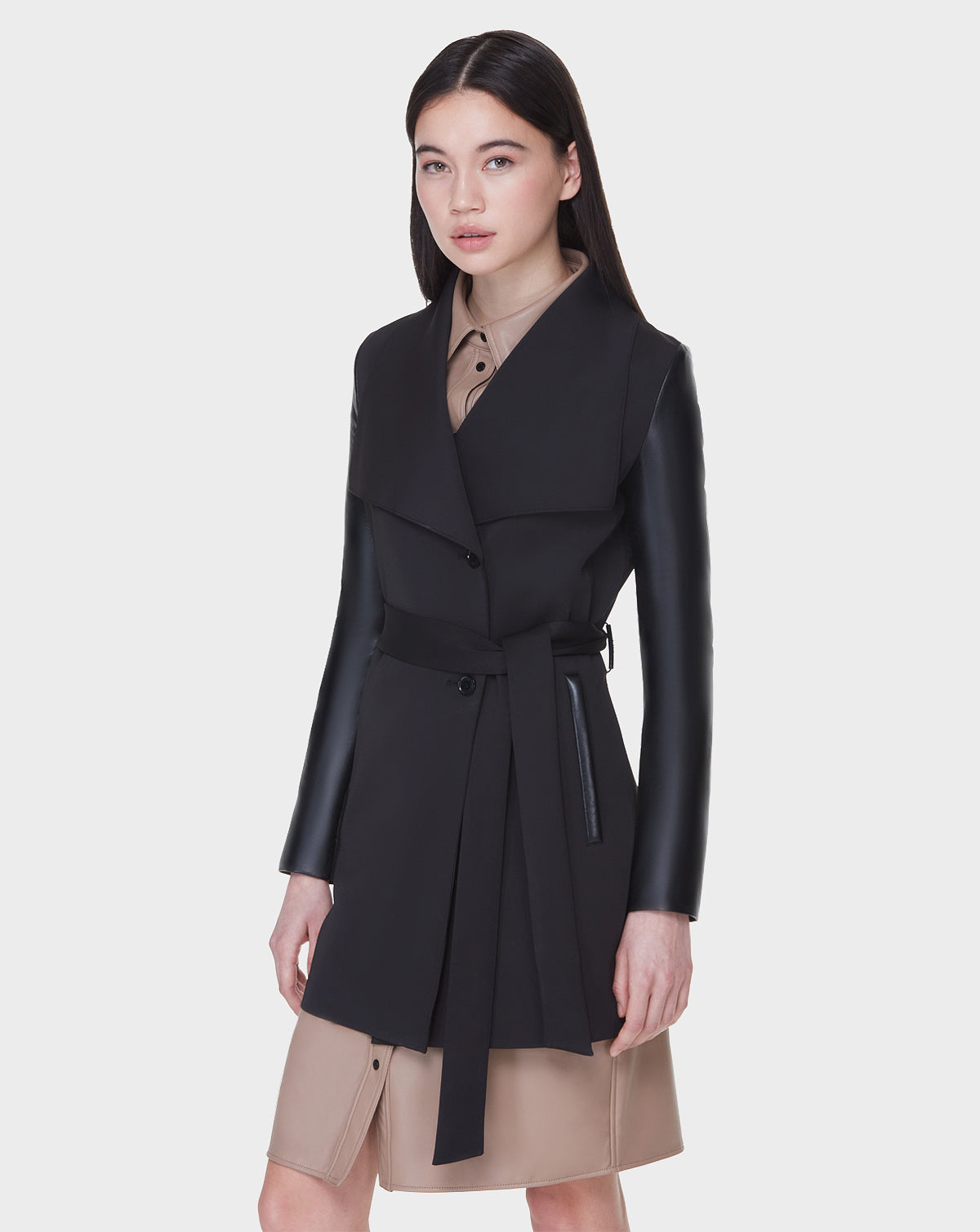 A woman with straight dark hair wears the Rudsak MELBA - L122007 BLACK women's coat with recycled leather sleeves over a tan shirt dress, standing against a plain white background.