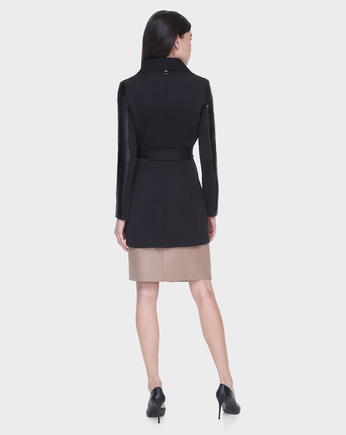 A woman with straight dark hair models the Rudsak MELBA - L122007 BLACK coat, featuring recycled leather sleeves and taupe bottom panels. She faces away, revealing the back details and black high heels against a plain light background.