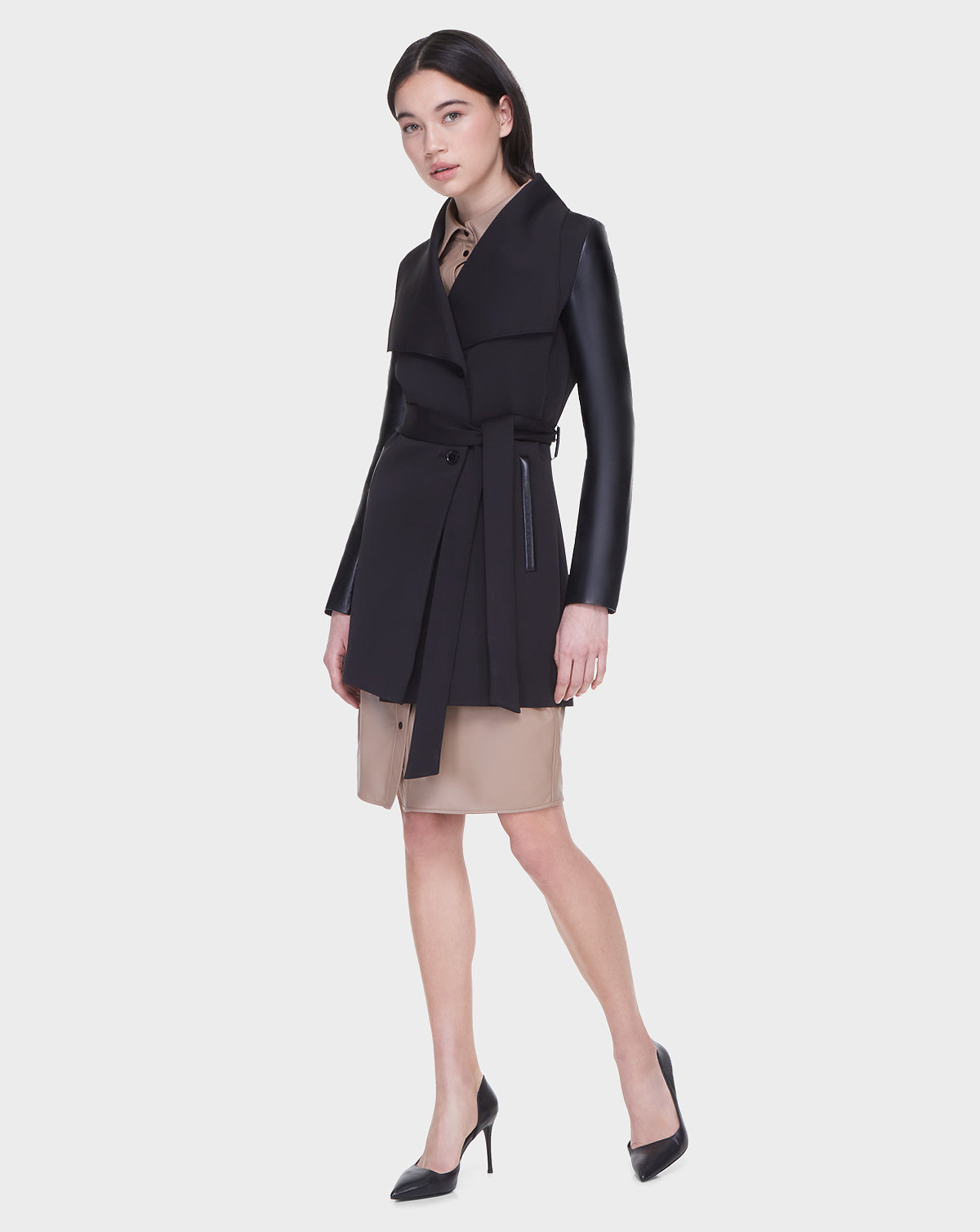 A woman poses against a white background wearing the Rudsak MELBA - L122007 BLACK coat, which features recycled leather sleeves, layered over a tan dress and black high heels.